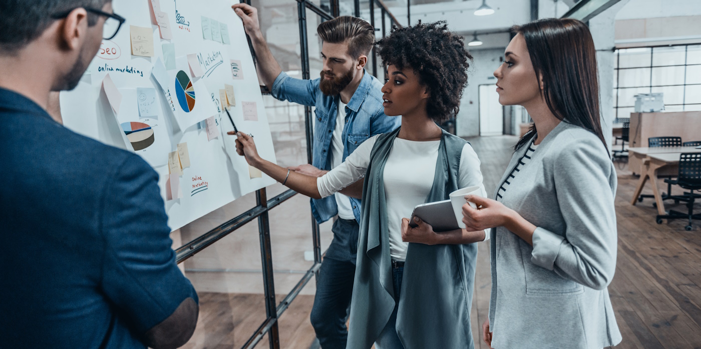 Group of young modern people in smart casual wear planning business strategy while young woman pointing at infographic displayed on the glass wall in the office